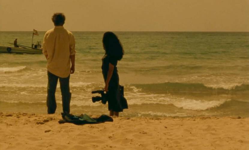 Ein Mann und eine Frau stehen am Strand und schauen auf das Meer, die Frau, rechts im Bild, hält eine Filmkamera in der Hand.