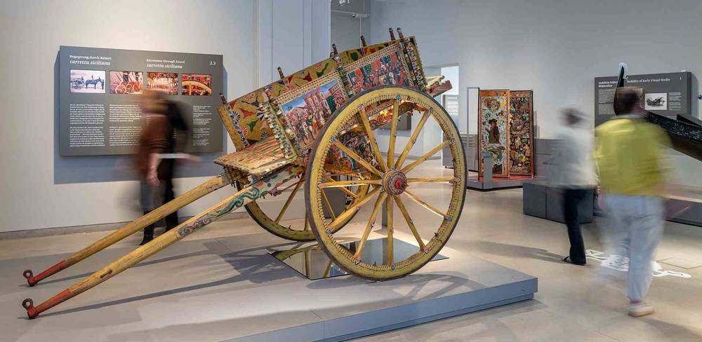 Ansicht der Dauerausstellung „Kulturkontakte. Leben in Europa“ – Bunt bemalter sizilianischer Karren im Museum, umgeben von unscharfen Personen. Informationstafel: "Interpreting Sicilian histories carros siciliani".