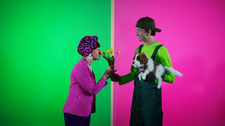 Ding Dong – Person mit pinkem Blazer und Kopftuch hält gelbe Tulpen, lacht mit person mit Hund. Grüne und pinke Hintergründe.