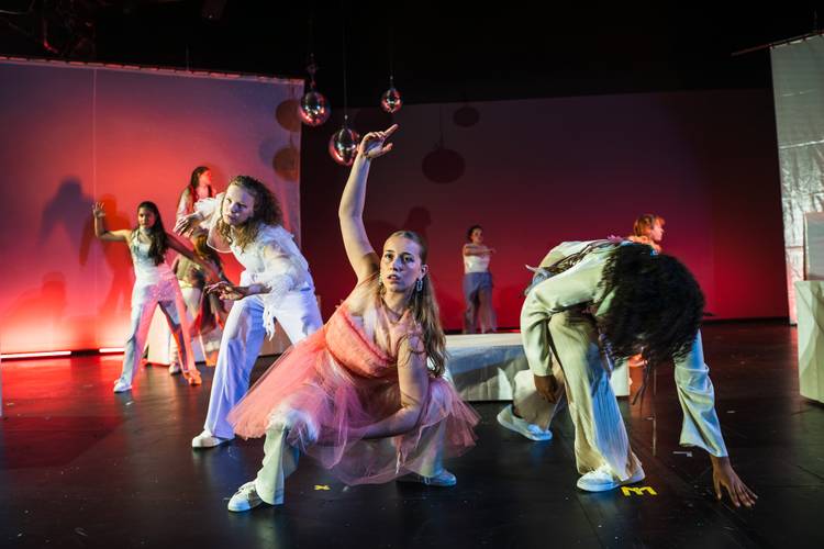 TRAUER//FALL, POLYLUX, Schauspiel Köln – Auf dem Bühnenfoto sind in etwas sieben Personen abgebildet. Die rechte seite des Bildes schimmert in einem grünen Licht und die linke in einem roten. Im Vordergrund ist eine weiblich gelesene Person die ein rosa Kleid trägt und hockt. Sie schaut nach vorne aber nicht in die Kamera, sie hat ihren Arm elegant über sich ausgestreckt. Links und rechts von ihr aber weiter hinten sind noch zwei Personen die ebenfalls in interressanten Positionen verharrt sind. Bis nach ganz hiinten der Bühne geht das weiter so.