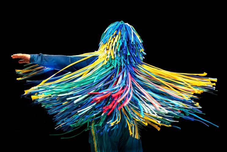 The Sticky Dance | (3-7) & (3-5) – Person mit buntem Fransenanzug in Bewegung, gelbe, blaue und rote Streifen wirbeln um den Körper, schwarzer Hintergrund.