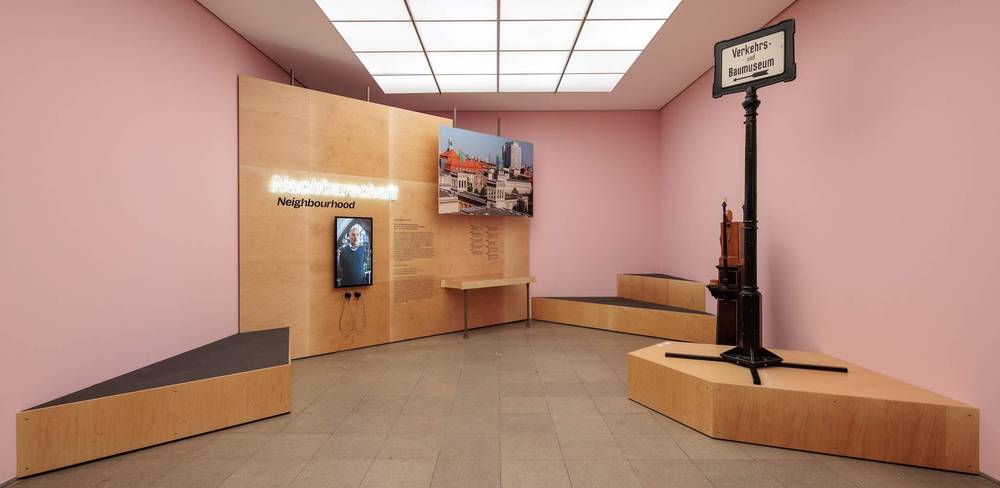 Ausstellungsansicht “Forum Hamburger Bahnhof“, Hamburger Bahnhof - Nationalgalerie der Gegenwart, 2023 – Raum mit rosa Waenden, Holzpodesten, einem Bild, einem Fernseher, Leuchtschrift "Nachbarschaft Neighbourhood" und Verkehrsschild "Verkehrsbaumuseum".