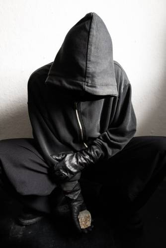 SAN PAOLO. Das Martyrium von Pasolini – Person in schwarzem Kapuzenpullover und Handschuhen hockt mit gesenktem Kopf. Gesicht nicht sichtbar. Hält einen Hammer senkrecht vor sich.