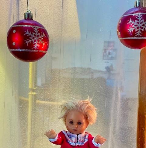 Puppe in rotem Kleid mit Spitze, umgeben von glitzernden roten Kugeln mit Schneeflockenmuster, vor einem verschwommenen Hintergrund.