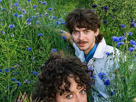 Notitzen an die Welt – Zwei Personen in einem Kornblumenfeld; vorne eine Person mit lockigen Haaren und rotem Lippenstift, hinten eine Person mit Schnurrbart in weißem Hemd.