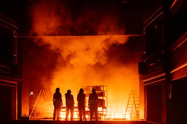 Vier Personen stehen vor oranger Rauchwand auf einer Buehne im Theater, flankiert von zwei Leitern.
