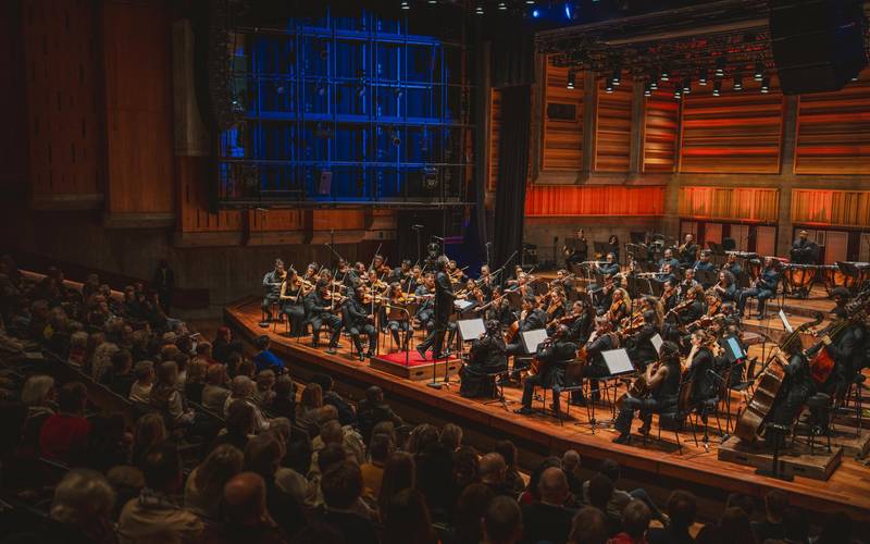Chineke! Orchestra – Orchester auf einer Holz-Bühne spielt vor Publikum. Dirigent steht in der Mitte, Musiker*innen mit Streichinstrumenten sitzen in Reihen.