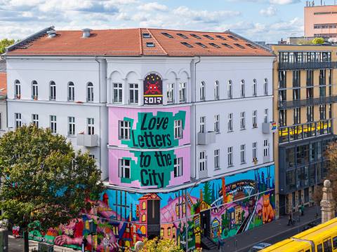  – Graffiti an der Hauswand des Museums mit Text: "Love letters to the City". Gelber Zug passiert im Vordergrund. Bäume im Herbstlaub. Urbaner Kontext, Tageslicht.