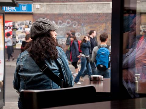 © Biene Pilavci – Eine Person mit langen Haaren sitzt auf einer Bank und schaut auf die Straße.