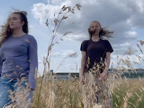 The field – Zwei Personen stehen auf einer Wiese mit hohem Gras. Beide haben lange Haare. Der Himmel ist bewölkt. Im Hintergrund ist ein Gebäude zu sehen.