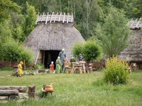 Die Darsteller:innen des Fördervereins Museumsdorf Düppel e.V. stellen mittelalterlichen Alltag nach. Besucher:innen können diesen live miterleben. – Ein Mann und mehrere Kinder in mittelalterlicher Kleidung halten sich vor einem rekonstruierten Haus auf dem Dorfplatz des Museumsdorf Düppel auf