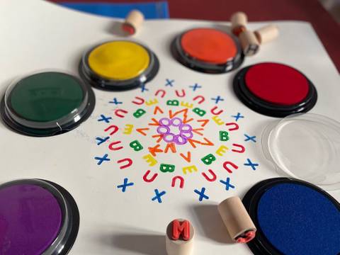 Mit Buchstabenstempeln und Farben entstehen bunte Muster. – Ein weißes Blatt mit farbig gestempelten Buchstaben sowie Stempelutensilien