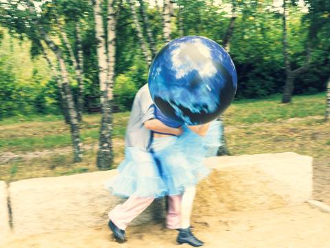  – Person trägt ein blaues Tutu und hält einen großen aufblasbaren Globus. Hintergrund mit Bäumen und Sandfläche.