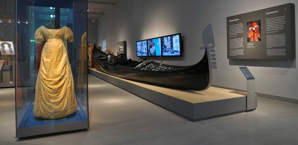 Ansicht der Dauerausstellung „Kulturkontakte. Leben in Europa“ – Eine Ausstellungsraum mit einer gelben Kleidvitrine und einer schwarzen Gondel. Rechts Texttafel: "Begegnungen", "Encounters". Wandbildschirm zeigt Kanäle.