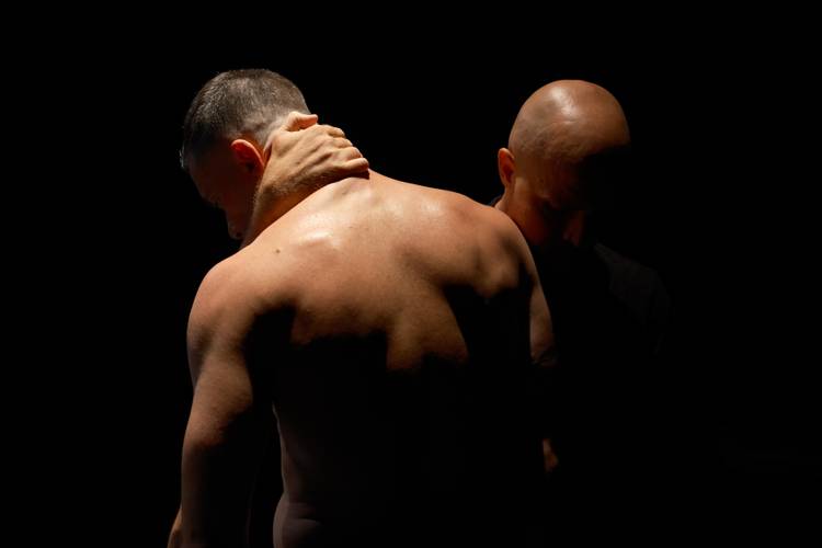 THE SHAPE OF A MAN – Zwei Personen im Dunkeln; eine mit freiem Oberkörper, schaut nach unten. Eine Hand berührt den Nacken der anderen Person, die sich teilweise im Schatten