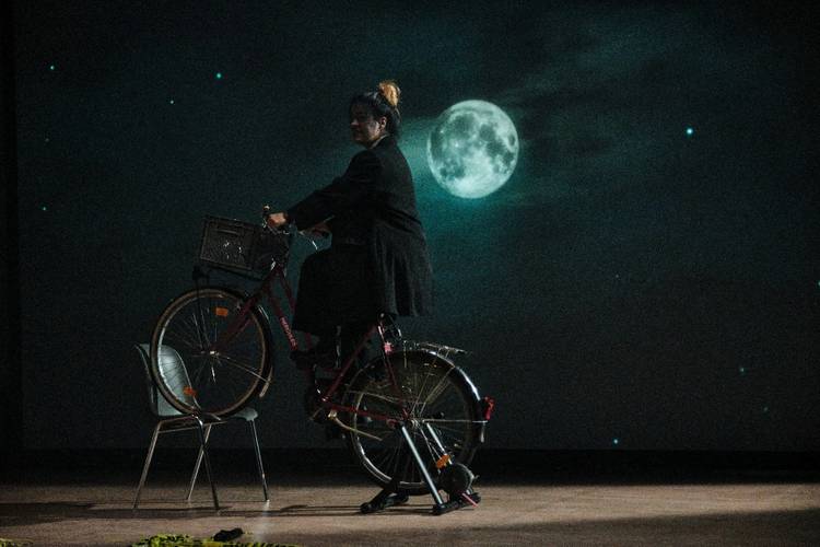 Person auf Fahrrad, das auf einem Stuhl steht, mit Mond und sternenklarem Himmel im Hintergrund. Sie trägt einen dunklen Mantel und hat hochgesteckte Haare.
