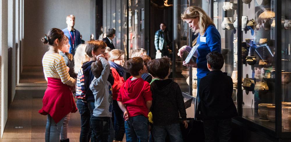 Familienworkshop im Neuen Museum – Person führt eine Gruppe von Kindern vor einer Vitrine voller Artefakte. Museumsausstellung mit Keramikobjekten. Helles Tageslicht beleuchtet die Szene.