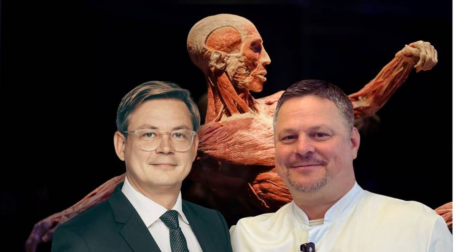 Körperwissen live: Ein Anatomieabenteuer über das Leben – Zwei lächelnde Personen vor einer menschlichen Anatomie-Ausstellung. Eine Person trägt Anzug, die andere ein weißes Hemd.