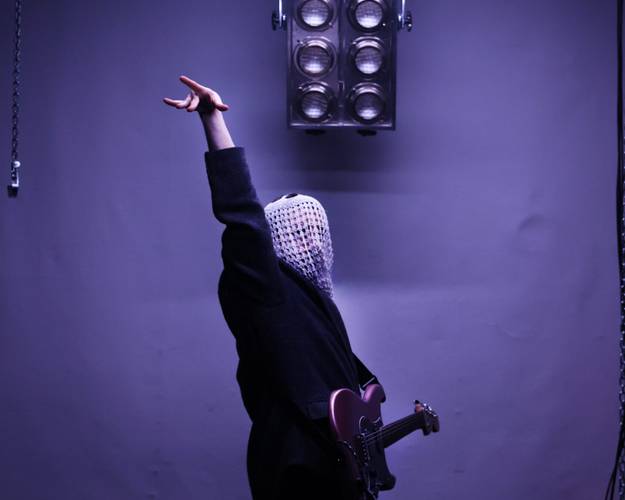 Eine Person mit gehäkeltem Tuch über dem Gesicht hält eine E-Gitarre und zeigt mit dem erhobenen Arm nach oben. Überhead ist eine große Lampe.