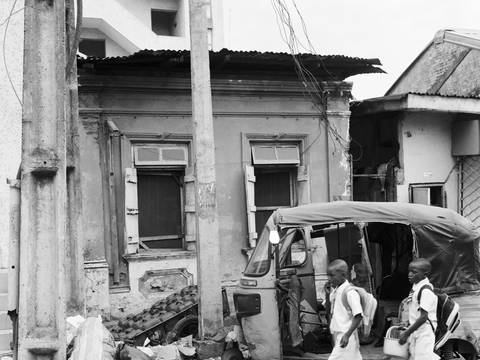 Akinbode Akinbiyi, Lagos Island, Lagos, 2016, Aus der Serie: „Lagos: All Roads“, seit den 1980er Jahren – Schwarz-weiß-Fotografie von zwei Schulkindern, die von rechts in das Bild laufen. Im Hintergrund sind das Gerüst eines kleinen Fahrzeugs und eine Hausfassade zu sehen.