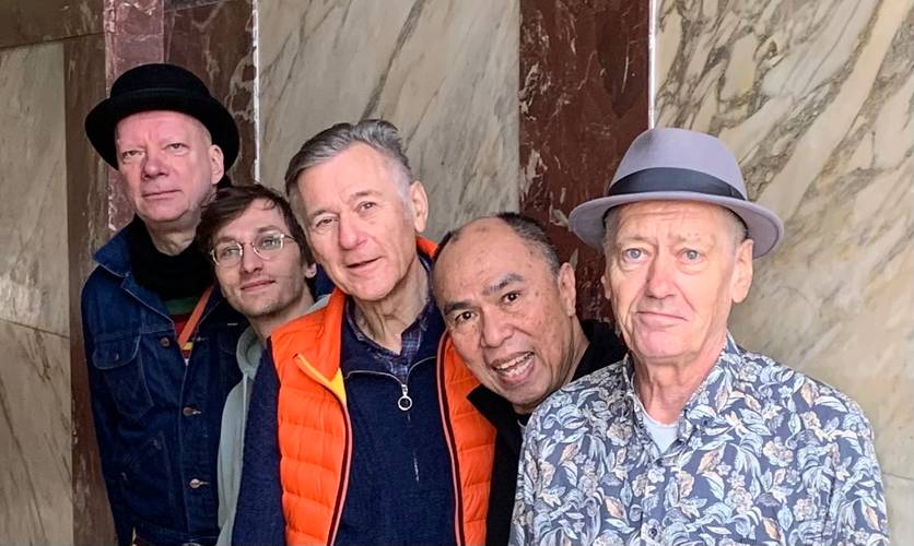 George Kranz & friends – Fünf Personen stehen nebeneinander, manche tragen Hüte und Brillen. Eine Person trägt eine orange Weste, eine andere ein Hemd mit Blumenmuster. Marmorsäulen im