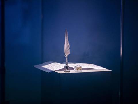  – Feder in Halter, Tintenfass und Papier auf Tisch in dunkler Umgebung, bläuliches Licht.