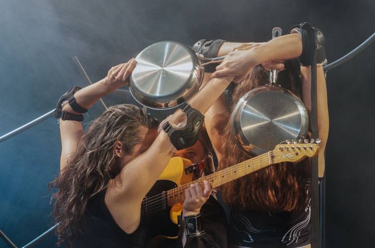 BANG! – Drei Personen auf einer Bühne, halten Kochtöpfe und Trommelstöcke. Eine Person spielt E-Gitarre. Bühnennebel im Hintergrund.