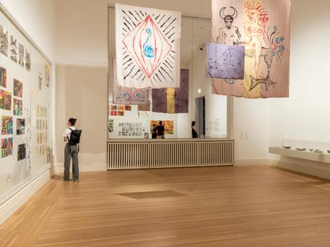 Gabriele Stötzer: "Dabei sein und nicht schweigen", Installationsansicht, Gropius Bau, 2026 – Besucher betrachtet moderne Kunstwerke von der Künstlerin Gabriele Stötzer in einer Galerie mit Holzfußboden. An den Wänden hängen abstrakte Zeichnungen und bunte Textilien mit symbolischen Motiven wie Augen, Spiralen und Figuren. Im Hintergrun
