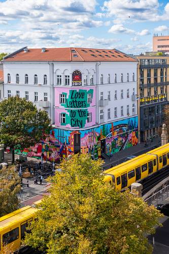 Graffiti an der Hauswand des Museums mit Text: "Love letters to the City". Gelber Zug passiert im Vordergrund. Bäume im Herbstlaub. Urbaner Kontext, Tageslicht.