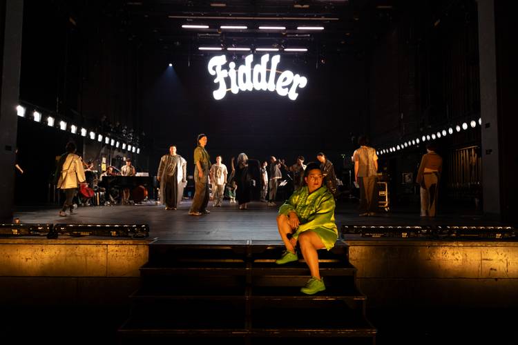 Bühne mit mehreren darstellenden Personen, Person in grünem Oberteil sitzt vorne, Leuchtschrift "Fiddler" im Hintergrund.