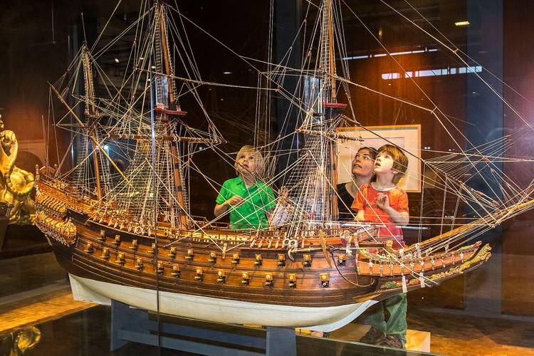 Die prachtvollen Schnitzereien der schwedischen VASA von 1628 zeigen, dass Schiffe auch als Repräsentationsobjekte und Ausdruck von Macht dienten. – Drei Kinder stehen auf einem Podest hinter einer Vitrine und betrachten ein großes, detailreiches Schiffsmodell. Hinter ihnen steht eine Frau.