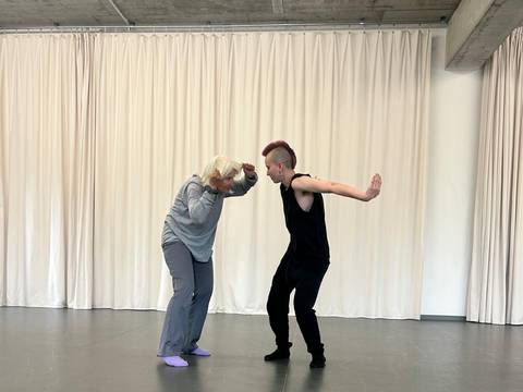  – Zwei Personen stehen gegenüber auf Tanzboden vor weißen Vorhängen, eine Person in Grau mit kurzen weißen Haaren, die andere in Schwarz mit kurzen roten Haaren.
