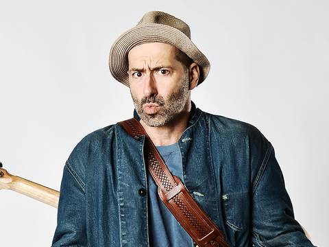 FIL – Mit neuer Show! – Person mit Strohhut und Jeansjacke, Gitarre auf dem Ruecken, schaut mit fragendem Gesichtsausdruck in die Kamera.
