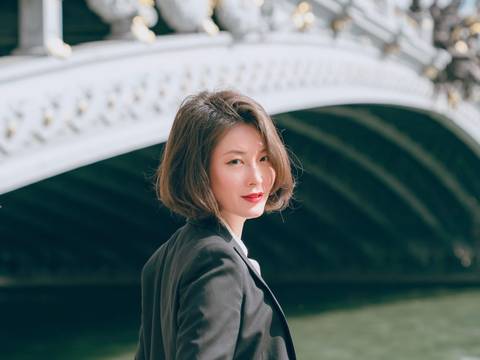 Shin Young Lee – Shin Young Lee mit kurzen braunen Haaren und einem schwarzen Blazer steht leicht lächelnd an einem Fluss, mit einer dekorativen Bogenbrücke im Hintergrund.