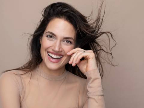 Marina Maximilian – Lachende, weiblich gelesene Person mit langen, wehenden Haaren und transparentem Oberteil vor neutralem Hintergrund.