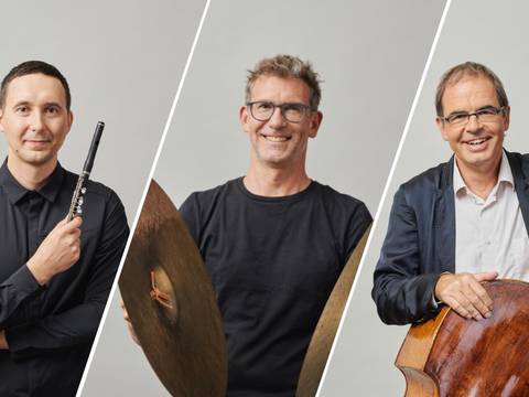 Jan Schlichte, Egor Egorkin und Sebastian Krunnies – Drei Personen stehen nebeneinander, eine mit Querfloete, eine mit Becken, eine mit Kontrabass. Studiofotografie mit neutralem Hintergrund.