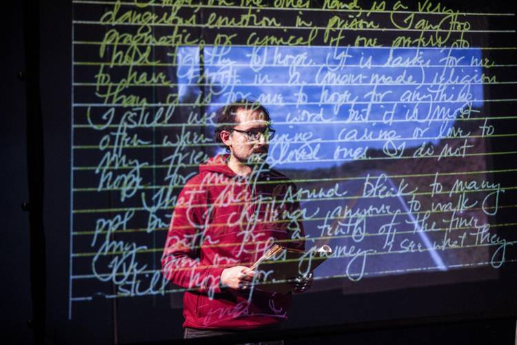 Person mit Brille und rotem Kapuzenpullover vor Projektionsleinwand mit handschriftlichem englischem Text.
