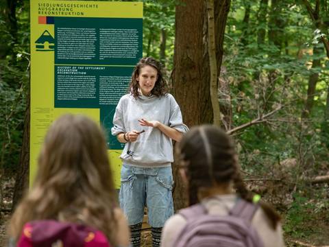 Die Führung informiert unter anderem über die Entstehung des heutigen Museumsdorfs. – Eine Frau erzählt vor einem Schild stehend die Geschichte der mittelalterlichen Siedlung im Museumsdorf Düppel