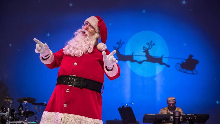 Das Märchen von der Prinzessin und dem Weihnachtsmann – Person in Weihnachtsmannkostüm vor einer blauen Kulisse mit Rentierschlitten-Silhouette. Im Hintergrund eine Person an einem Keyboard.