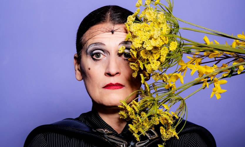 La Signora – Person vor lila Hintergrund haelt gelbe Blumen. Gesicht teils bedeckt, Make-up mit schwarzem Eyeliner, rote Lippen, kurze dunkle Haare. Kleidung schwarz.