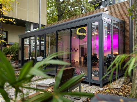 Einblicke Gartenhaus – Moderner Pavillon mit Glaswaenden, innen pink beleuchtet, mit Aufschrift "Einblicke" auf runder Tafel, umgeben von Baeumen und Pflanzen.