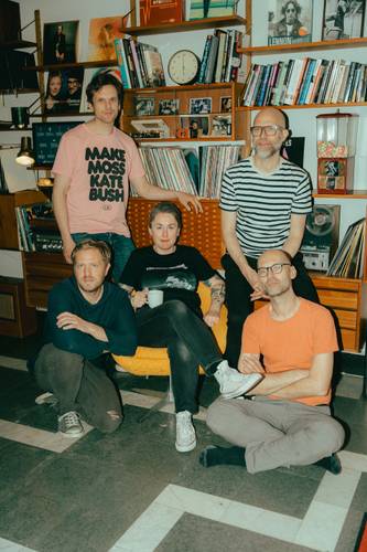 Elin Forkelid – Gruppe von fünf Personen in einem Wohnzimmer mit Bücherregalen, Schallplatten und einem auffälligen T-Shirt in Rosa.