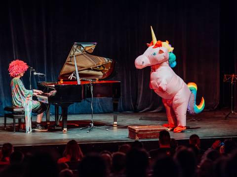 Karneval der Tiere – Person mit bunter Perücke spielt Klavier auf Bühne. Neben der Person steht ein großes, buntes Einhornkostüm. Publikum im Vordergrund.