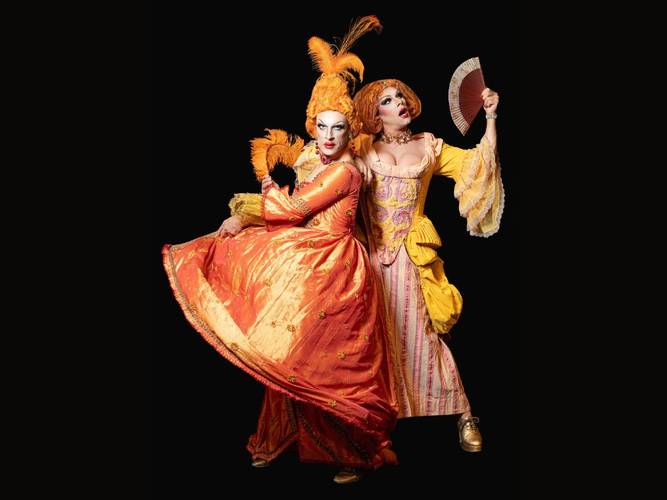 PINK GRIMM: Shakesqueer – God save the Queens – Zwei Personen in opulenten barocken Kostümen; links in orange mit Federhut, rechts in gelb-rosa mit Fächer. Beide mit stark geschminkten Gesichtern.