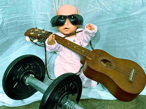  – Puppe mit Sonnenbrille haelt Gitarre, sitzt neben Hantel auf Stoffhintergrund.