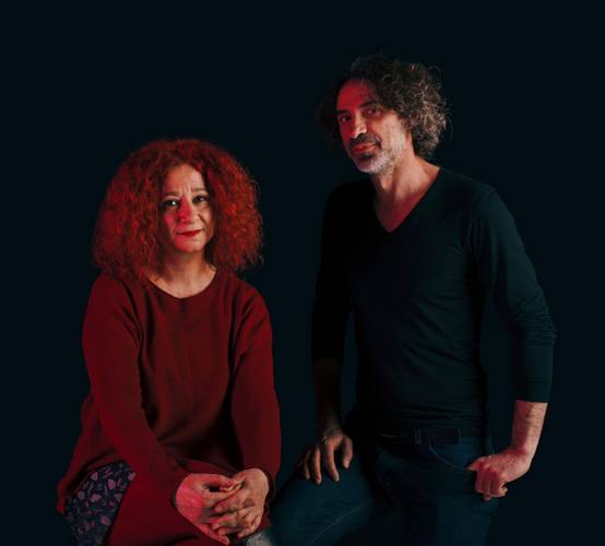 Das Künstler*innen Duo Lina Majdalanie und Rabih Mroué – Zwei Personen sitzen bzw. stehen vor dunklem Hintergrund eine Person in rotem Oberteil, die andere in dunkler Kleidung.