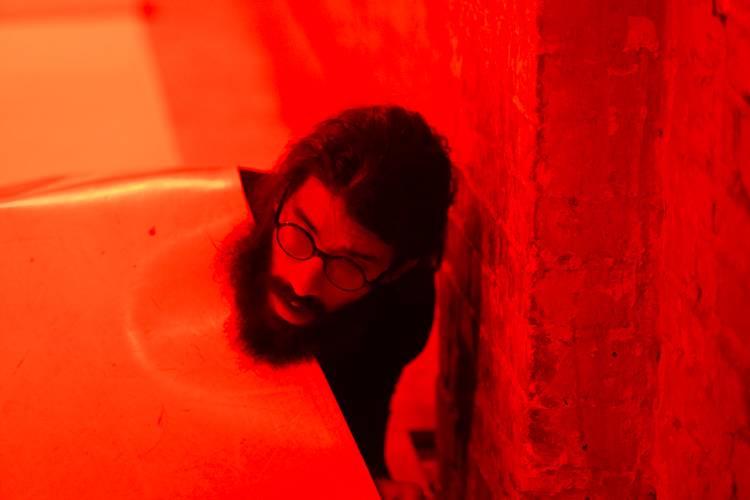 Person mit Brille und Bart schaut hinter einer Wand hervor, in rotes Licht getaucht.