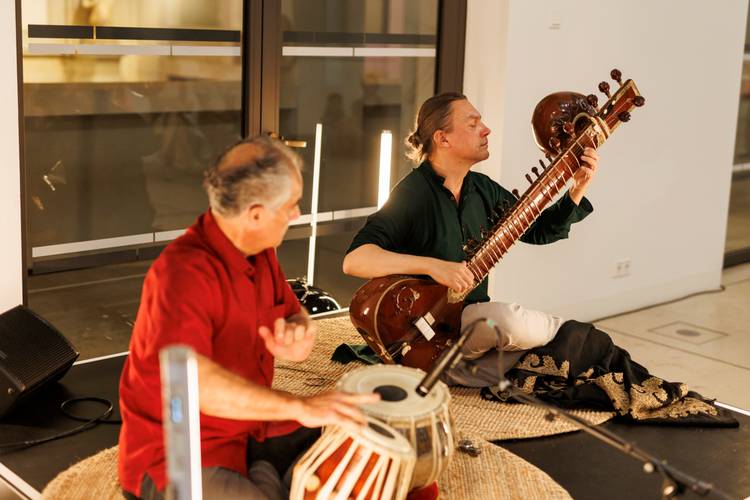Nacht-Ragas auf Sitar und Tabla bei der Lange Nacht der Museen 2024 im Humboldt Forum – Zwei Personen spielen Instrumente auf Teppich; eine Sitar, eine Tabla. Beide sitzen vor großen Fenstern, eine trägt eine rote, die andere eine grüne Kleidung.