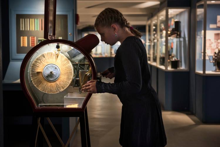 Am Mutoskop (um 1900) entsteht beim schnellen Drehen der Eindruck eines bewegten Films. – Ein Mädchen schaut in einen beleuchteten Kasten und dreht an einer Kurbel. Im Hintergrund sind Vitrinen mit Objekten zu sehen.
