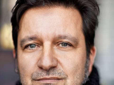 Julian Voloj – Person mit dunklen Haaren und Dreitagebart, trägt einen schwarzen Strickpullover, schaut ernst. Hintergrund unscharf.